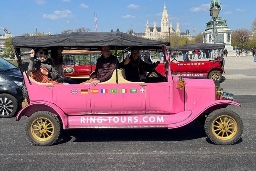 En pink veteranbil med passagerer, parkeret i et offentligt område med en historisk bygning i baggrunden. 5