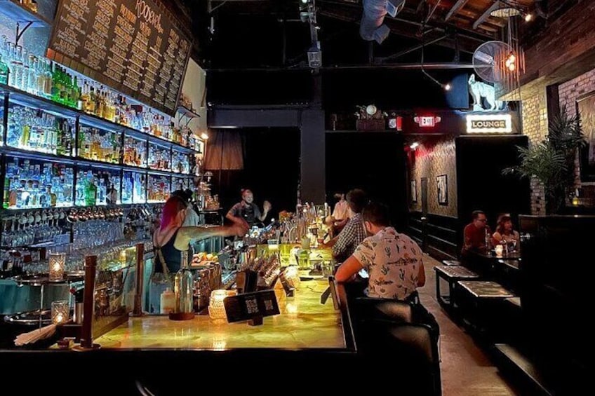 Een bar met flessenrekken, een zithoek en mensen die zitten en staan. 4
