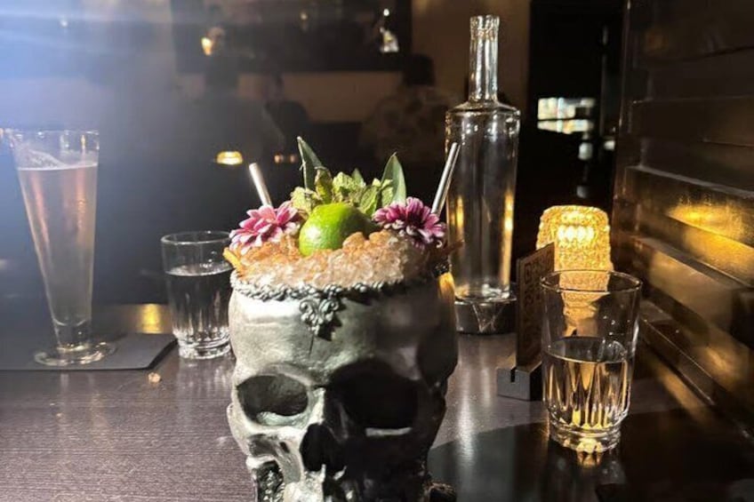 Een schedel-vormig drankje met een limoen en bloemen, omringd door glazen en een kaars. 3