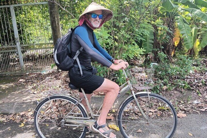 En person som cyklar på en cykel med en korg, bär en hatt, shorts och sandaler, och befinner sig i en naturlig miljö med träd och ett staket. 4