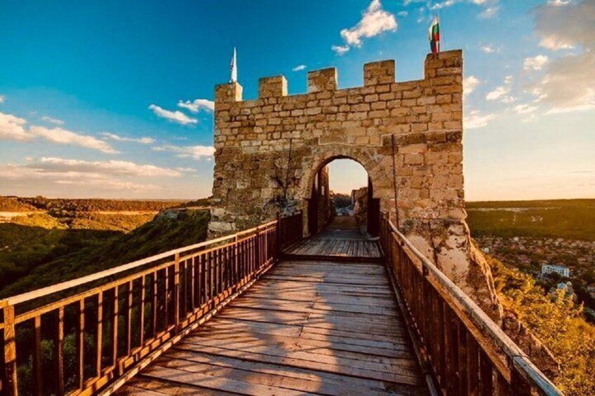 A stone archway leading to a castle, surrounded by a wooden bridge and a hilly landscape. 1