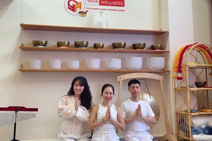 Three people in white clothing performing a yoga pose in a room with shelves of bowls and a decorative chair. 3