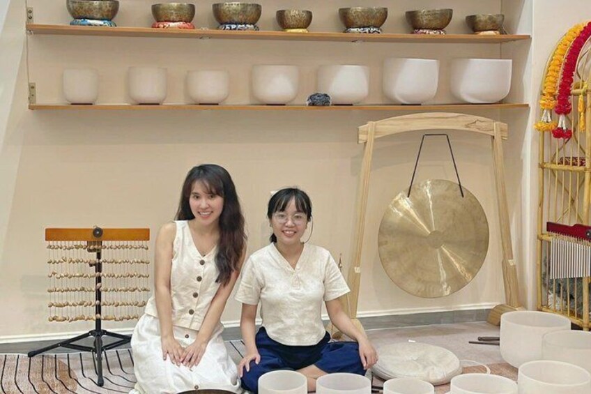 Two people sitting on the floor with bowls and a large gong in a room with shelves of bowls and a hanging gong. 2