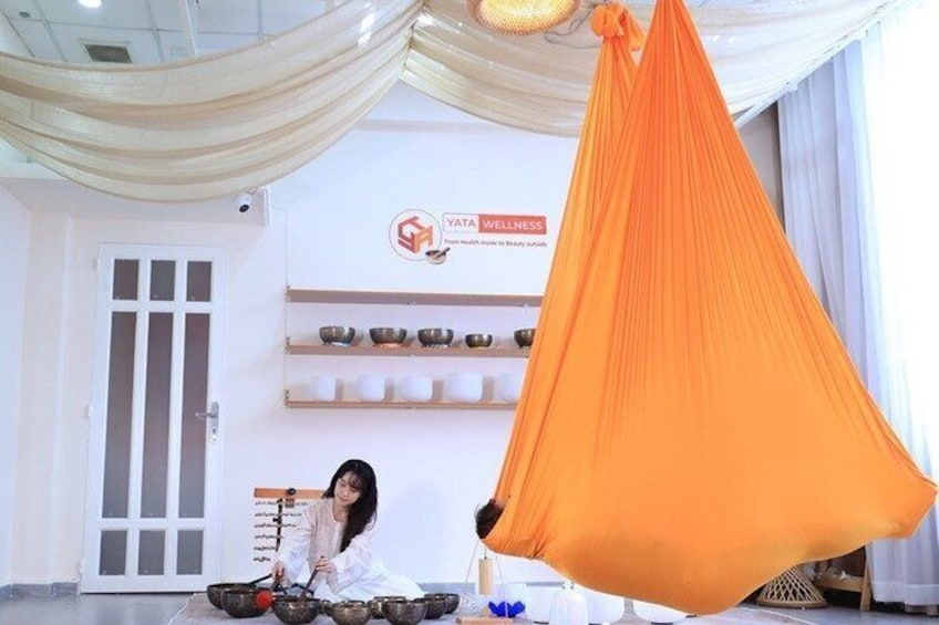 A person sitting on the floor with a hammock. There are shelves with bowls and a sign that says 'YATTA WELLNESS'. 1