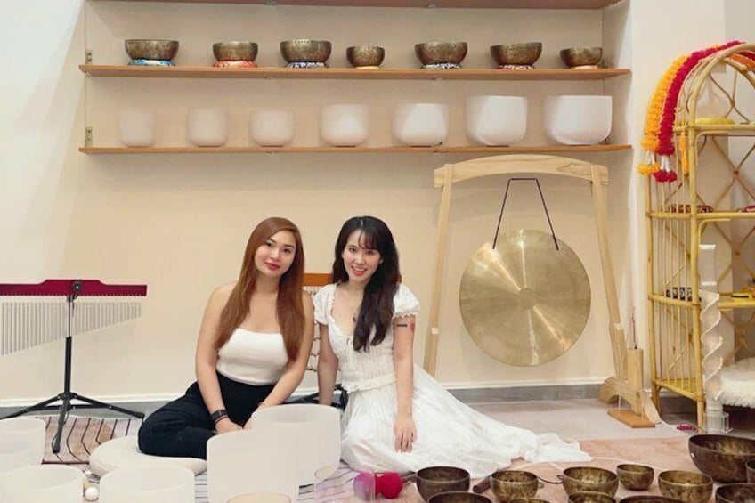 Two women sitting on a floor with bowls and a gong in a room with shelves of bowls and a stand with a red cloth. 4