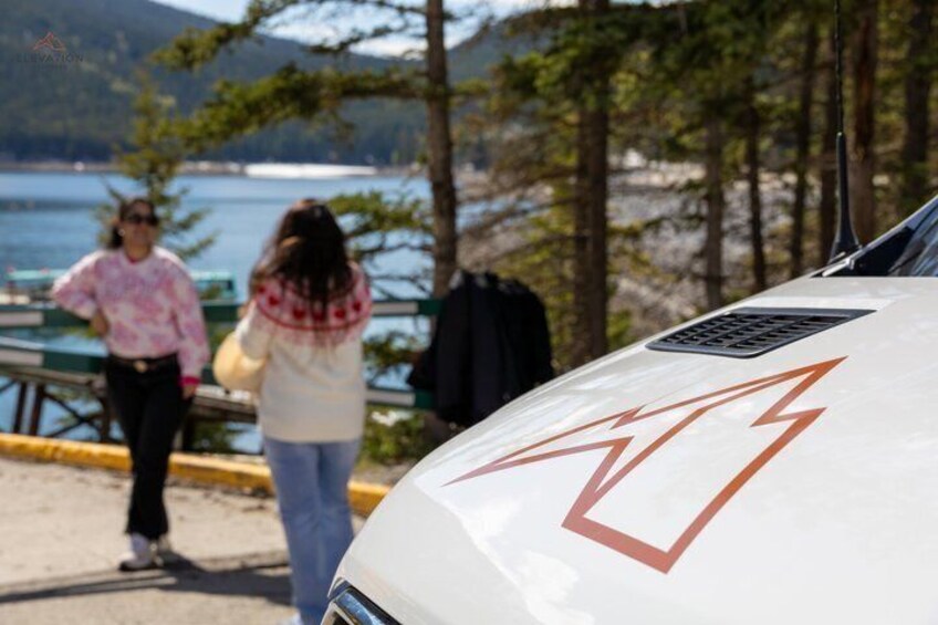 A white vehicle with a red arrow symbol on its hood, parked near a body of water with people in the background. 5