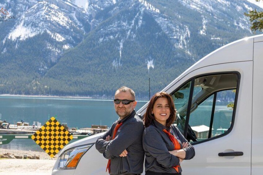 A couple standing beside a white van with a mountainous lake in the background. 4