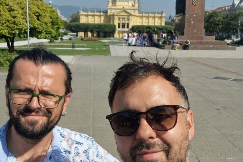 Two people in a public square with a historic building and a clock tower in the background. 2