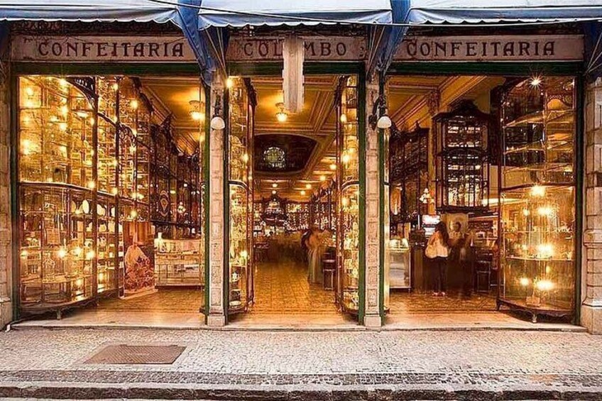 A brightly lit storefront with glass display windows and the word 'CONFEITARIA' above the entrance. 3