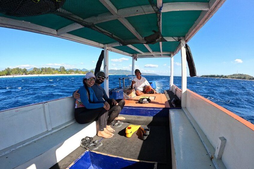 A boat with two people sitting on the deck, a blue container, and a person standing at the back. 3