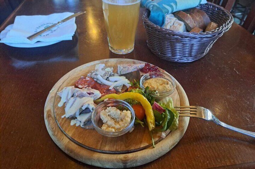 A wooden board with various meats, vegetables, and dips, accompanied by a glass of beer and a basket of bread on a wooden table. 5
