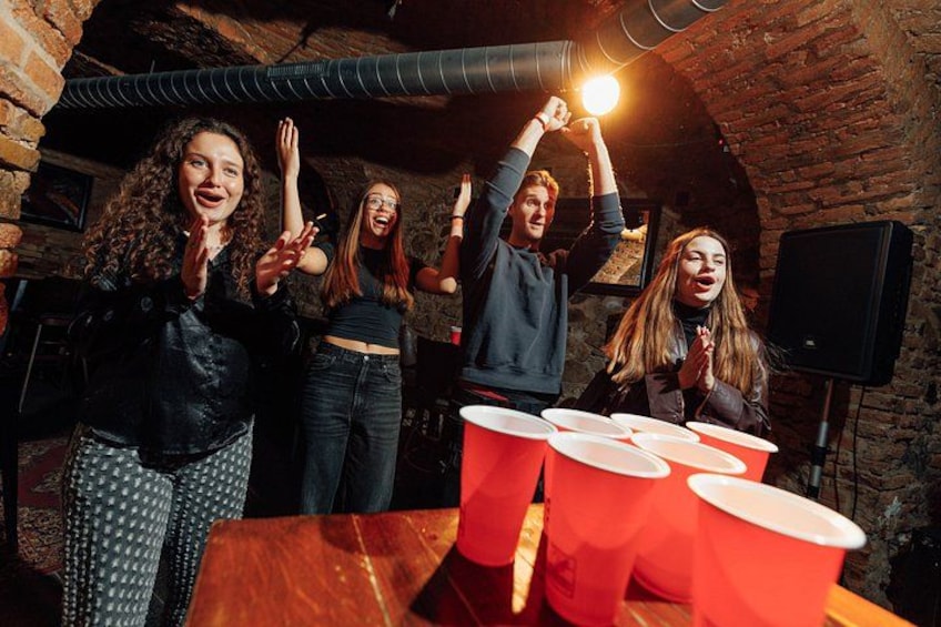 A group of people playing beer pong in a rustic room with brick walls and a wooden table. 3