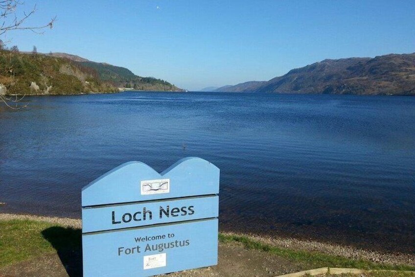 A sign reading 'Loch Ness Welcome to Fort Augustus' by a lake. 3