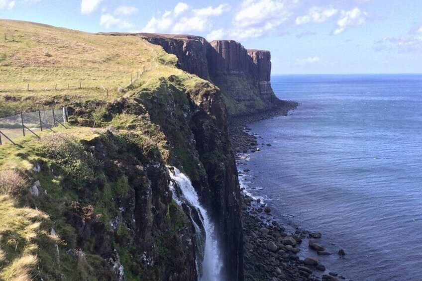 A coastal cliff with a waterfall, grassy terrain, and a clear sky. 1