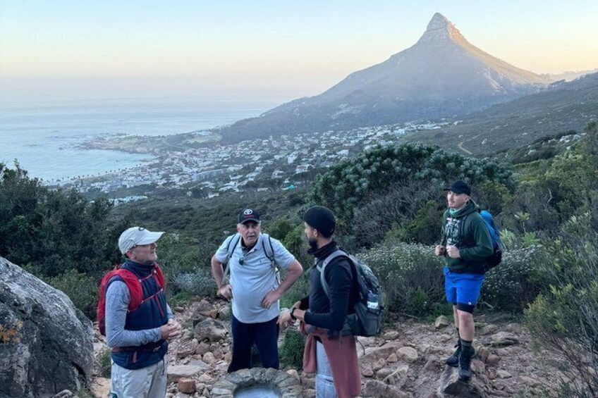 A group of hikers on a rocky trail with a view of a mountain and a coastal town in the background. 1