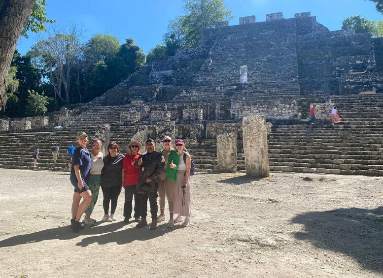 A group of people standing in front of ancient stone ruins with a large tree and a clear blue sky in the background. 1