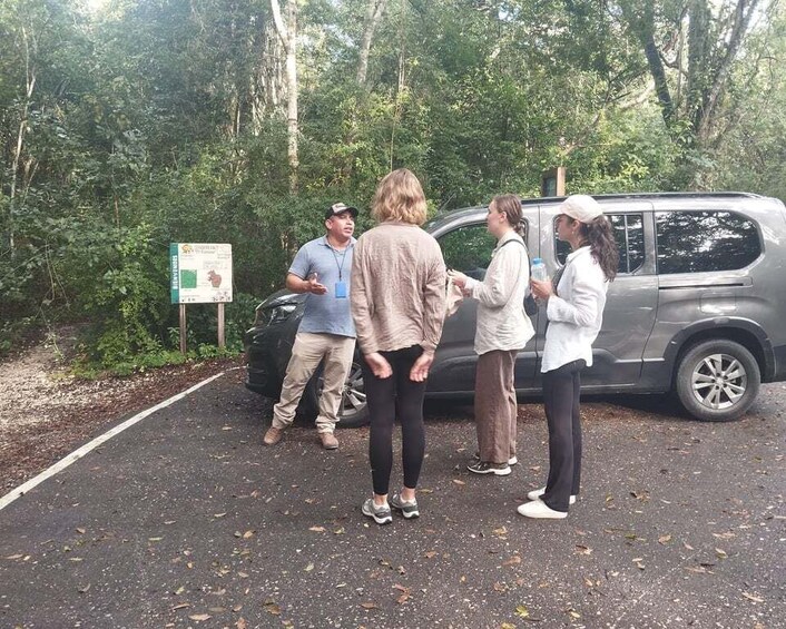 A group of people standing near a vehicle in a forested area with a signboard. 2