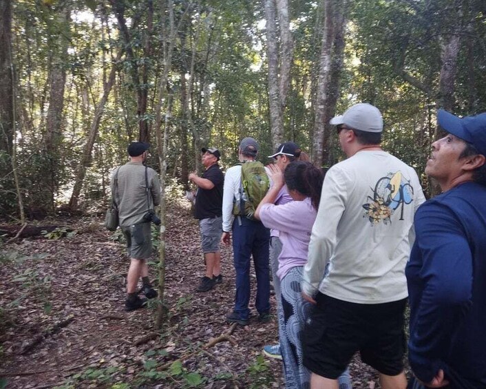 A group of people in a forested area, some standing and some sitting, with trees and a forest floor covered in leaves. 4