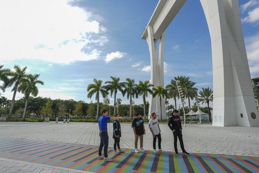 Un grupo de personas de pie sobre una alfombra a rayas de colores en una plaza abierta con palmeras y una gran estructura arqueada blanca al fondo. 4