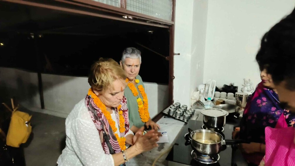 Two people wearing garlands, a stove with a pot, and kitchen utensils. 5