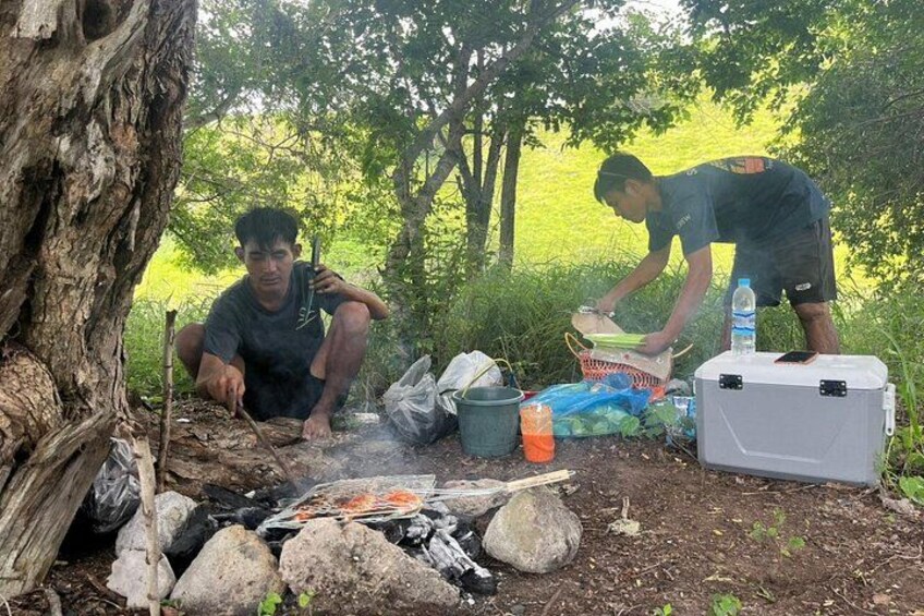 Two people cooking outdoors with a fire, surrounded by rocks and greenery. 5