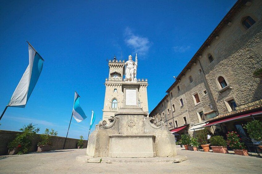 A historical monument with a statue, surrounded by stone buildings and flags. 3