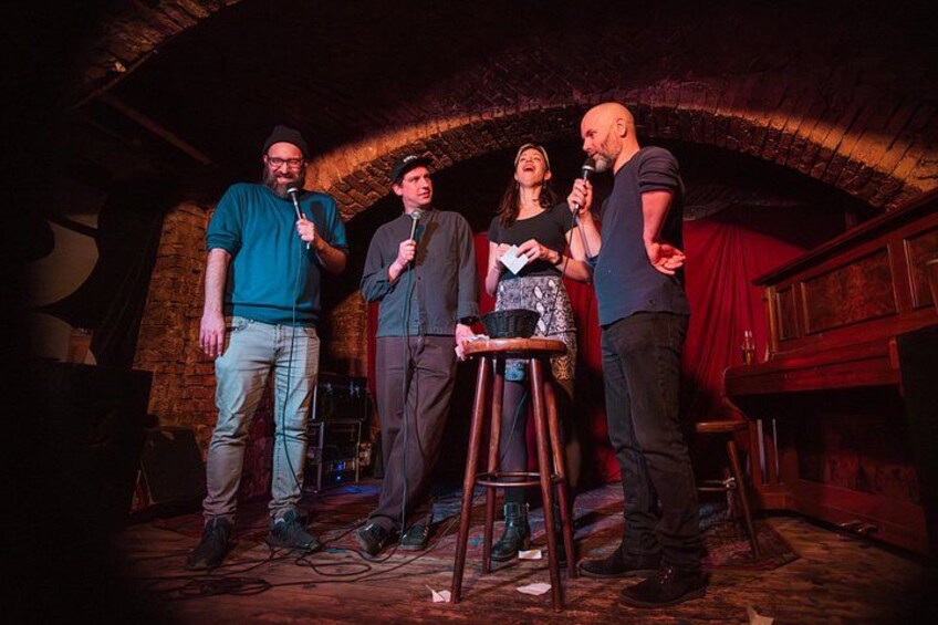 Un grupo de cuatro personas en un escenario, con un soporte para micrófono, un piano y un muro de ladrillos al fondo. 4
