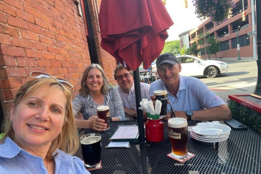 A group of people sitting at an outdoor table with drinks, a brick wall, and a street in the background. 1
