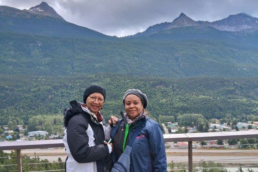 Two people standing on a balcony with a mountainous landscape in the background. 4