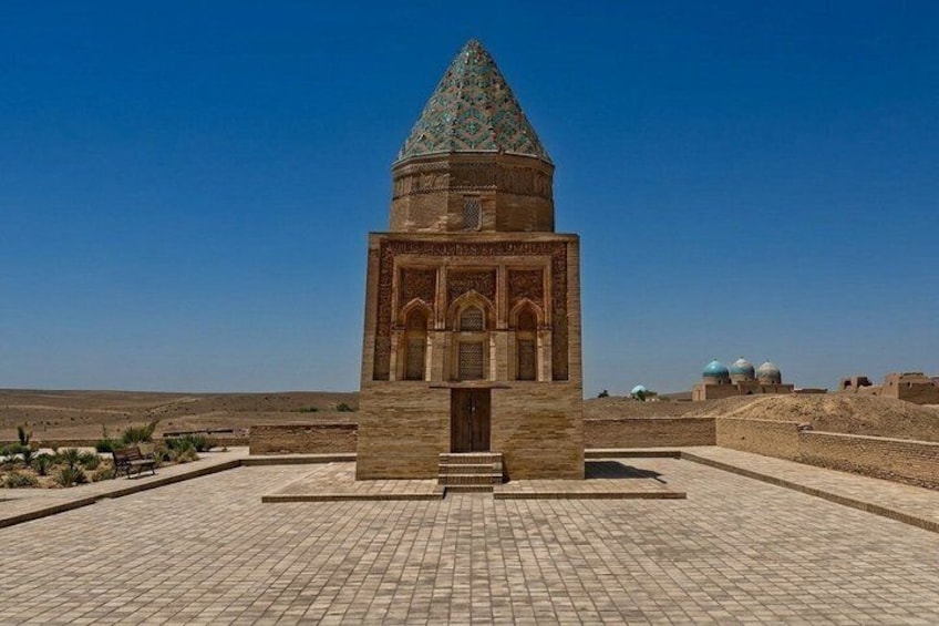 A historical monument with a pointed roof and intricate carvings, surrounded by a paved area and distant domed structures. 5