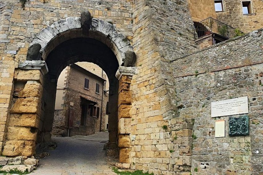 An old stone archway leading to a narrow street, with a building and a sign on the right. 4