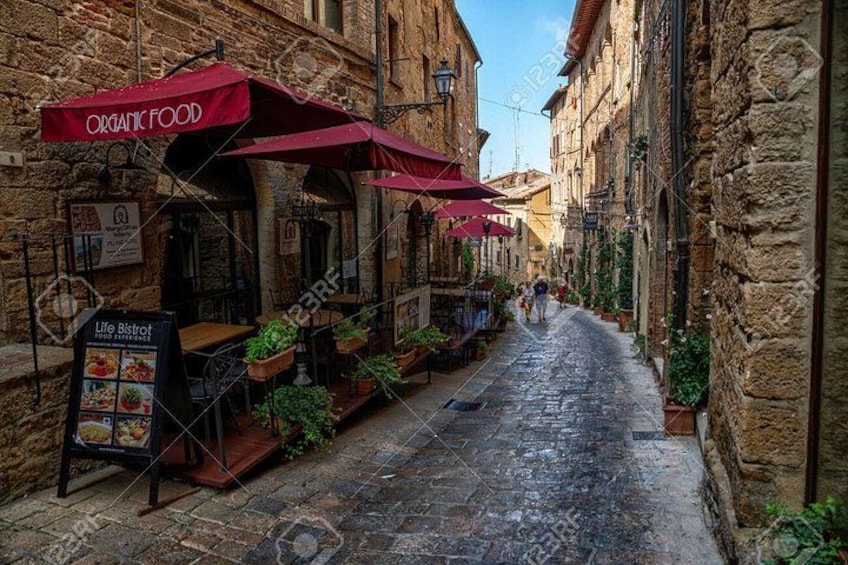 Una estrecha calle adoquinada con mesas al aire libre bajo sombrillas rojas, edificios de piedra y un letrero de Life Bistro. 3