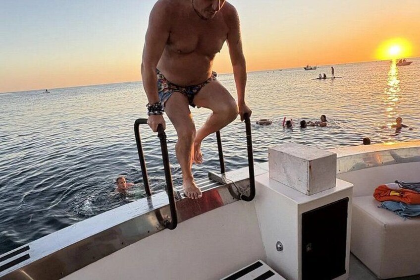 Un uomo in costume da bagno su una barca, aggrappato a una ringhiera, con l'oceano e il tramonto sullo sfondo. 3