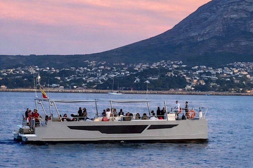 A boat with passengers on a calm body of water, a mountainous landscape in the background, and a distant shoreline with buildings. 4