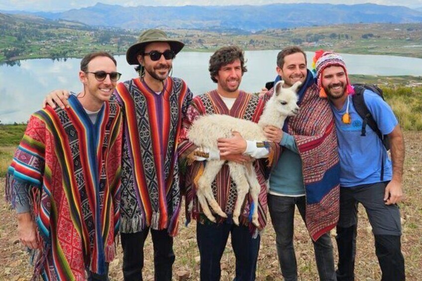 A group of people in traditional attire with a llama, standing by a lake with mountains in the background. 4