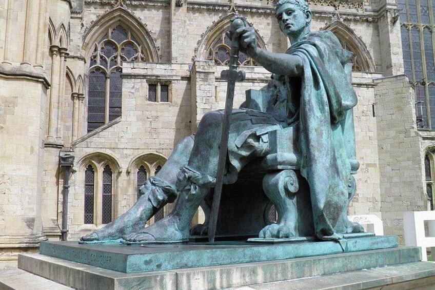 A bronze statue of a seated figure with a staff, set against a Gothic-style building with pointed arches and detailed stonework. 1