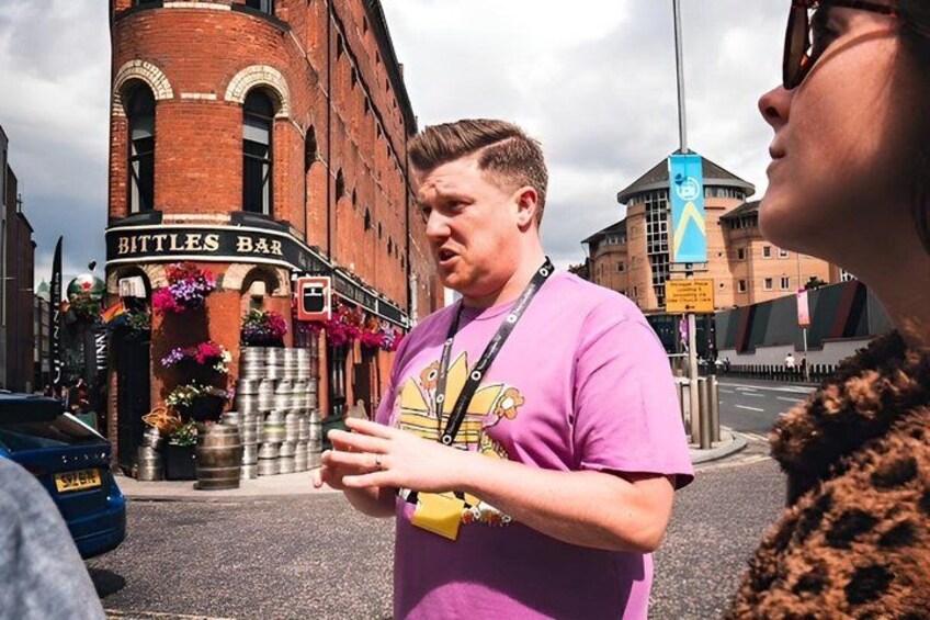 A man in a purple shirt standing on a street with a bar named 'Bittles Bar' in the background. 4