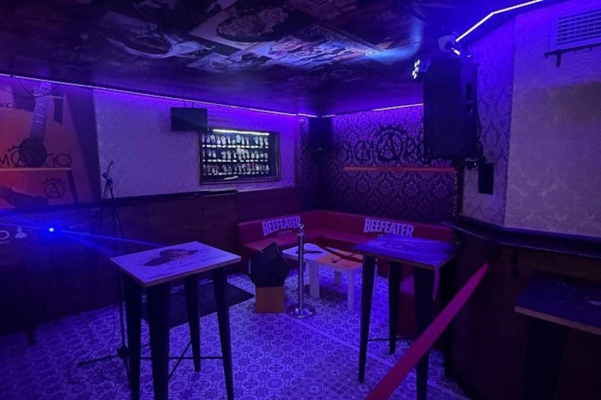 A dimly lit bar with tables and chairs, a wall-mounted TV, and a patterned floor. 5