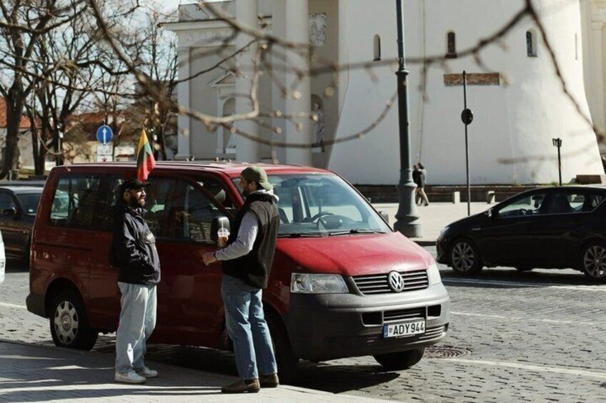 A red van parked on a cobblestone street with a flag on top, a building in the background, and a pedestrian walking by. 2