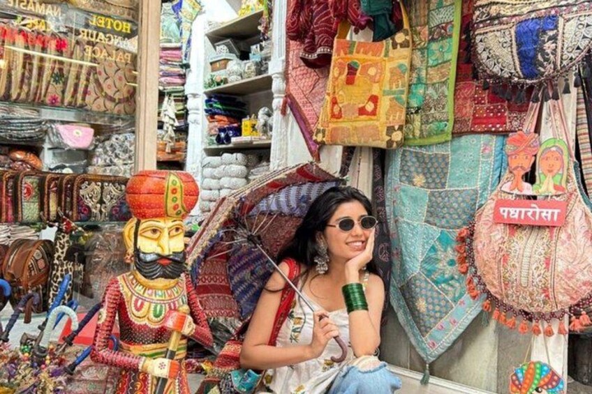 A person sitting in front of a shop with colorful textiles and bags. 1