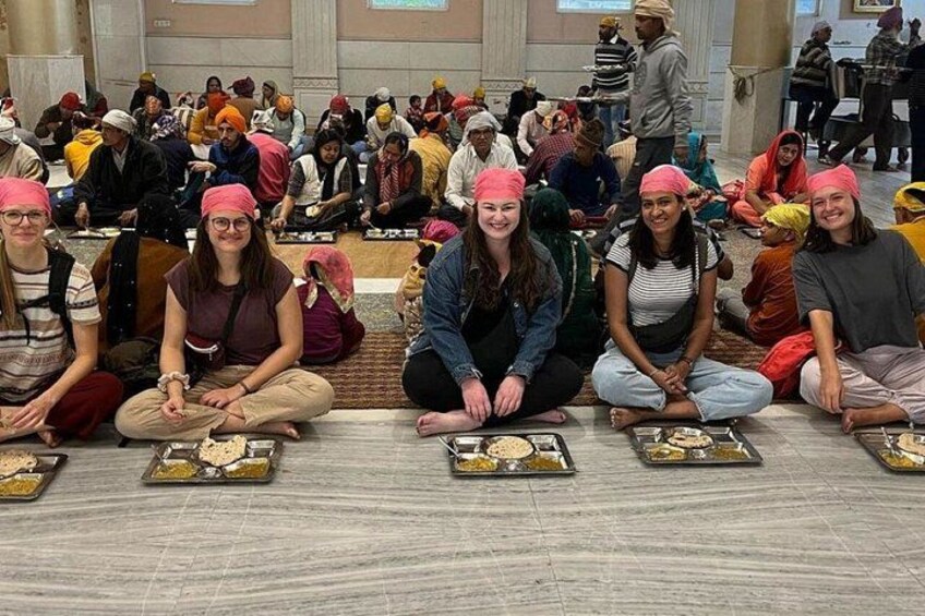 A group of people sitting on the floor in a large hall, some wearing pink headscarves, with trays of food in front of them. 5