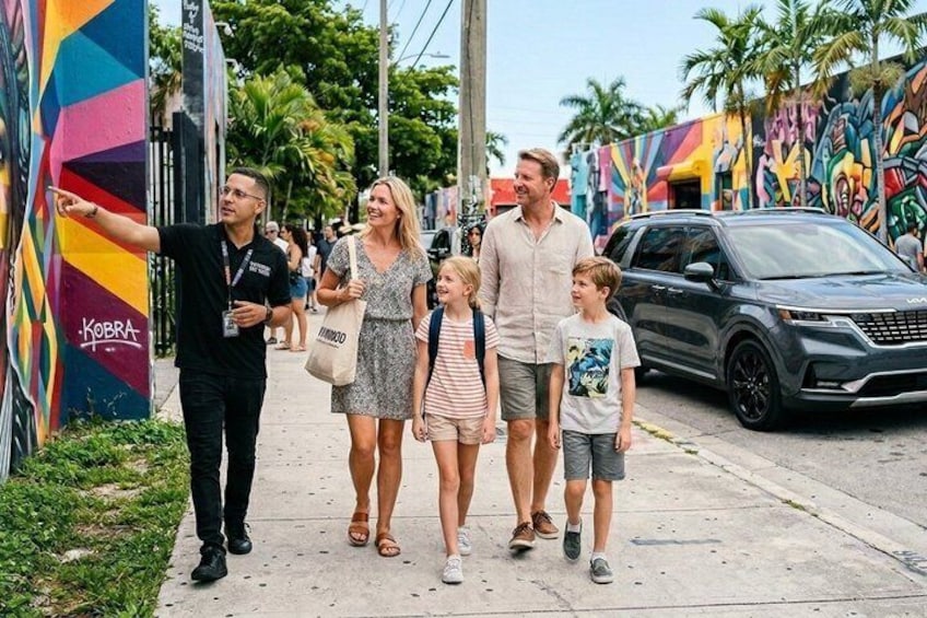 A family walking on a sidewalk with colorful murals and a parked SUV. 1