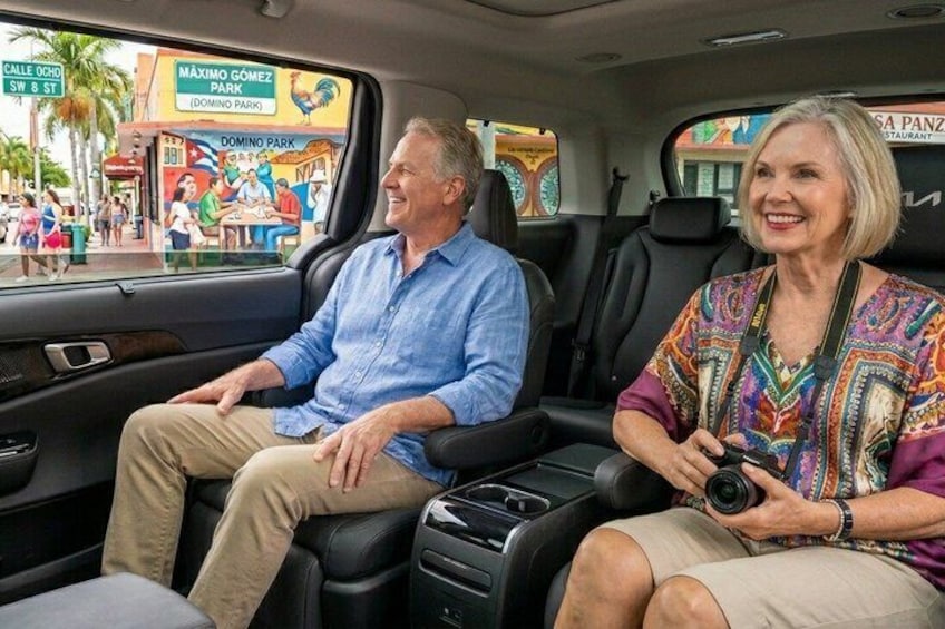 Two people seated in a car with a view of a street scene including a sign for 'Domino Park.' 2