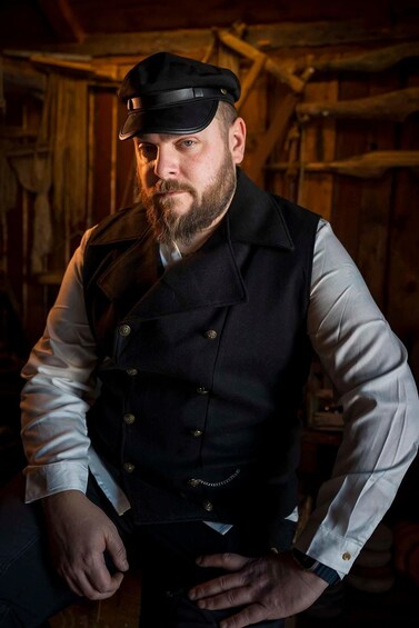 A person wearing a black cap, a dark vest, and a light-colored shirt, seated indoors with wooden walls and a rustic setting. 1