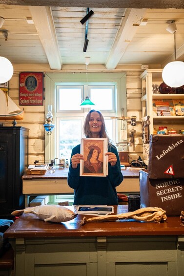 A person holding a framed picture in a rustic interior with wooden beams, a window, and shelves with various items. 5