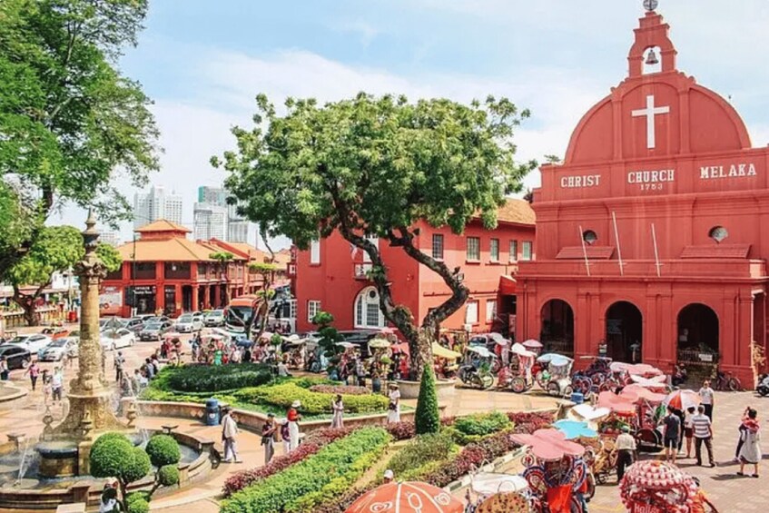 A bustling square with a prominent red church labeled 'Christ Church Melaka,' surrounded by greenery and a busy street with pedestrians and vehicles. 1