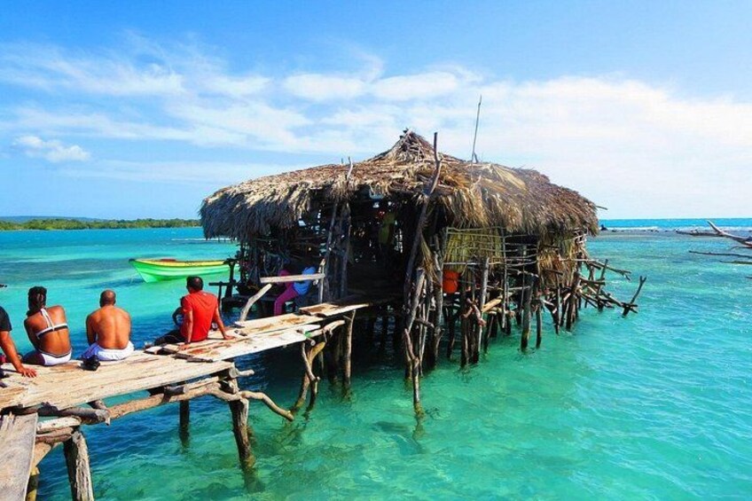 A thatched-roof hut on stilts over turquoise water, a wooden dock, and people sitting on the dock. 3