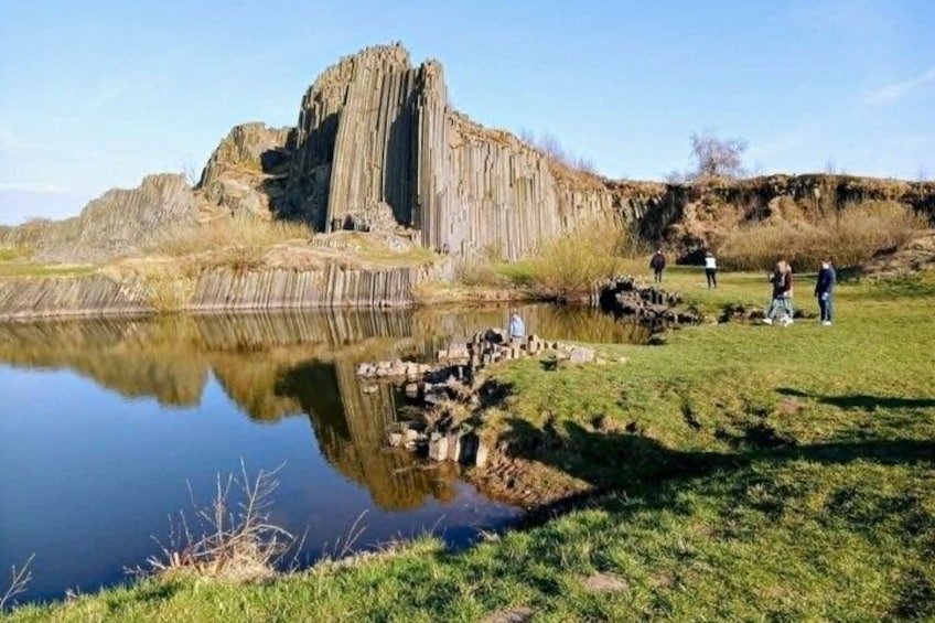 A rocky cliff by a calm lake with people walking on the grass. 1