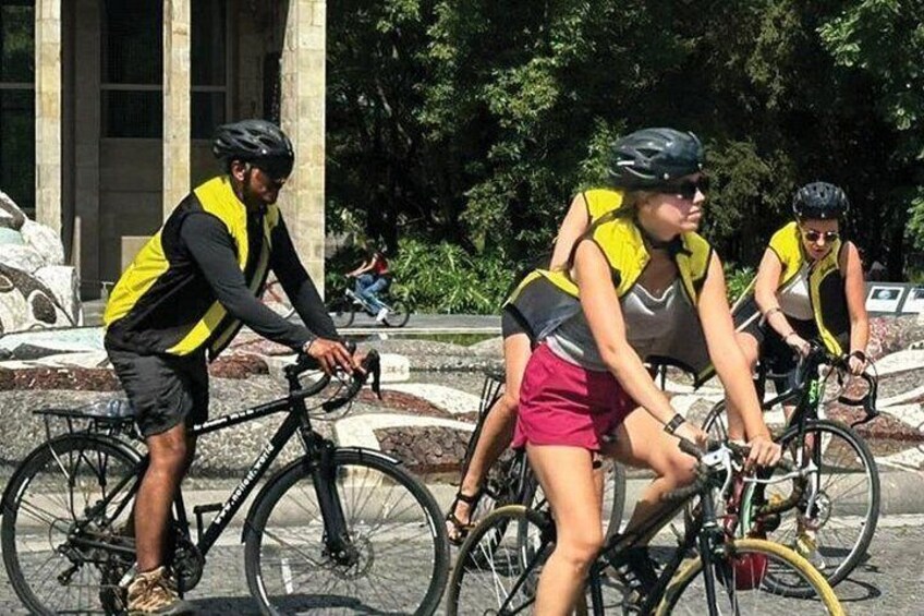 Three cyclists wearing yellow and black helmets and reflective vests, riding on a paved path with a stone border. 2