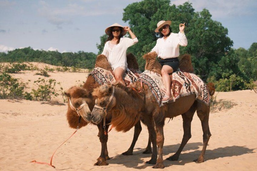 Two people riding camels in a desert with sparse vegetation and a clear sky. 2